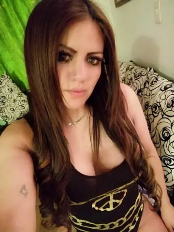 Woman with long brown hair wearing a black tank top sitting on a patterned floral sofa in a cozy room.