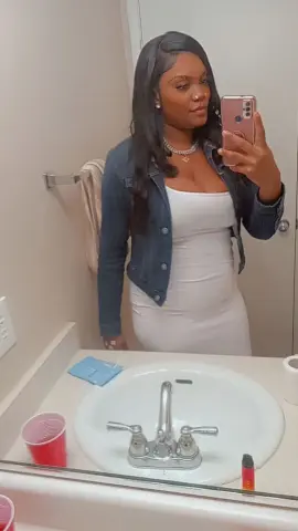 Young woman in a white dress and denim jacket taking a mirror selfie in a bright bathroom.