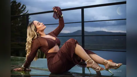 Elegant woman in a flowing brown dress relaxing on a balcony overlooking a misty mountain lake at twilight.