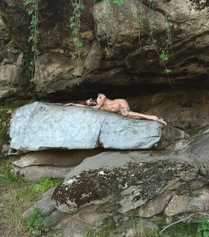 AlisaMoss lies naked on a large rock under a rocky overhang in a forest setting.