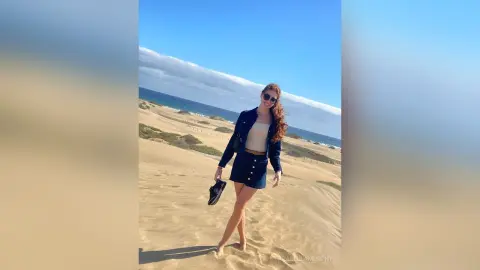 Stylish woman in a navy blazer and denim shorts walking on a sunny sand dune beach.