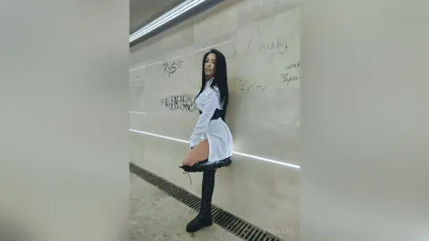 KymSkyler poses in a white shirt and black boots against a stone wall with graffiti, showing off her long dark hair and legs.