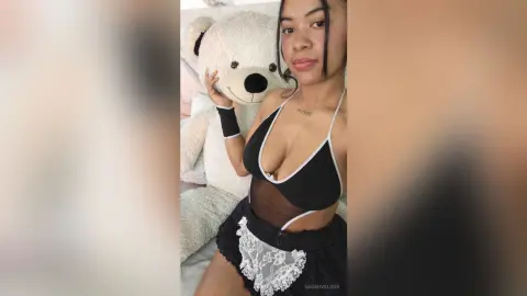 NaomiVelour poses in a black and white lace lingerie set next to a large white teddy bear, showing off her cleavage and slim waist.