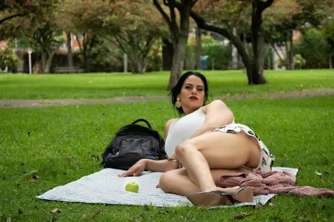 Woman in white tank top relaxing on a picnic blanket in a lush green park on a sunny day.
