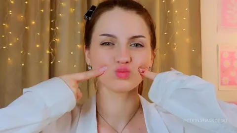 PetrinaFranceski pouts her lips and touches her cheeks, wearing a white shirt with her hair pulled back and string lights in the background.