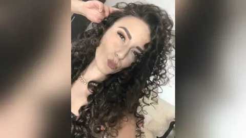 MontseFerrera pouts her lips while posing with her long, dark curly hair and a gold chain necklace.