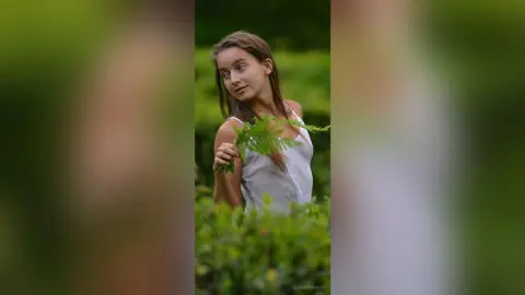 Young woman in a white tank top holding a green fern leaf in a lush, dreamy garden setting.