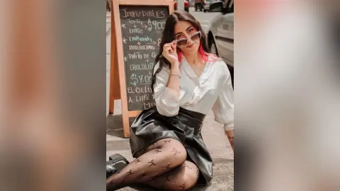 Young woman in white blouse and black leather skirt sitting by a cafe menu board in a sunny urban setting.