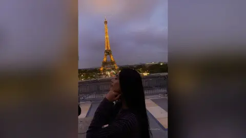 LucyMartin poses with her hand near her chin against a backdrop of the illuminated Eiffel Tower at dusk.