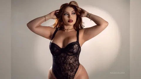 Glamorous brunette woman in black lace bodysuit posing confidently against a soft studio background.