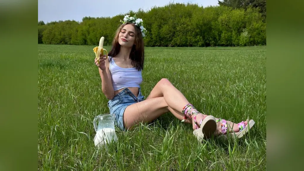 Young woman in white crop top and denim shorts eating a banana in a sunny green meadow.