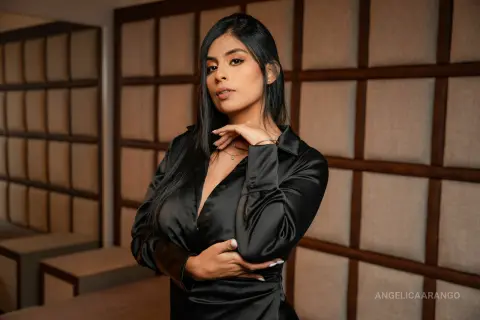 AngelicaArango poses in a black satin long-sleeve shirt with her hands near her neck and chest against a padded wall background.