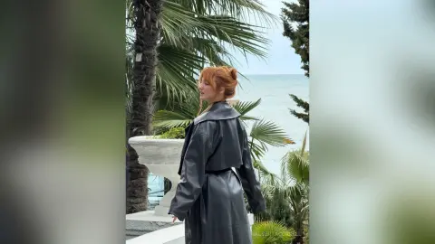 AlbertaCorrell stands outdoors in a black leather coat with her red hair styled up, looking out toward the ocean near palm trees.