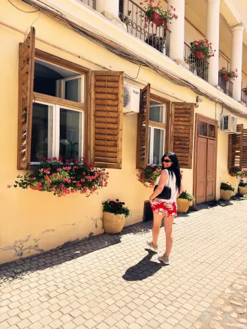 Woman in a white top and red floral skirt walking past a yellow building with flower boxes on a sunny day.