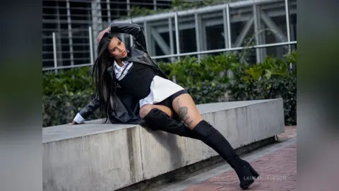 Stylish woman in a black leather jacket and over-the-knee boots leaning on a concrete ledge outdoors.