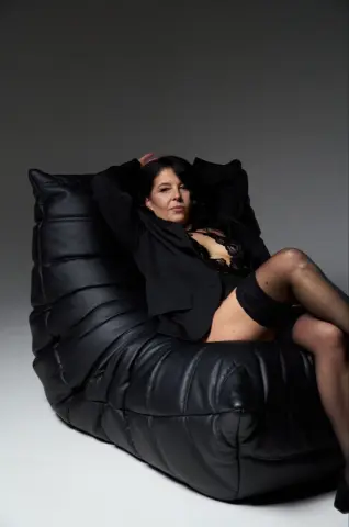 Elegant woman in black lingerie and blazer relaxing on a large black leather lounge chair in a studio setting.