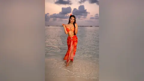 Young woman in an orange bikini and sarong walking through shallow ocean water at sunset.