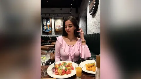 Young woman in a pink ruffled blouse enjoying a meal at a cozy restaurant with appetizers and drinks.