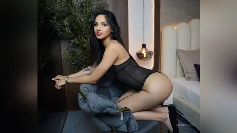 Smiling woman in black lace lingerie sitting on a grey chair in a modern bedroom setting.