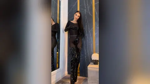 LeyaGrifin poses in a black sheer bodysuit and thigh-high patent leather boots against a blue and gold wall.
