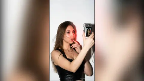 Young woman in a black leather top taking a selfie with an instant camera against a white background.