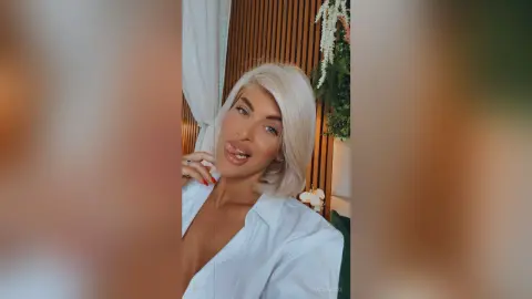 Blonde woman in a white button-down shirt posing with a playful expression in a warm, modern interior.