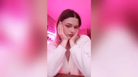 Young woman in a white ribbed sweater posing thoughtfully against a soft pink aesthetic background.