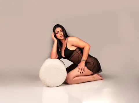 Curvy woman in a black sheer bodysuit posing elegantly on a white ottoman against a neutral studio background.