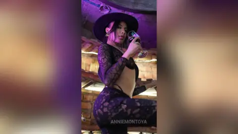 Stylish woman in black lace outfit and wide-brimmed hat sipping wine in a moody, dimly lit lounge.