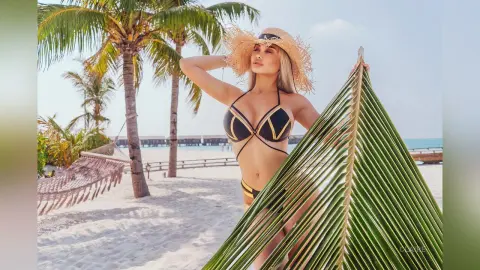 Blonde woman in a black bikini posing on a sunny tropical beach with palm trees and a hammock.