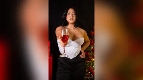 Elegant woman in a white corset and black trousers holding a glass of rosé in a festive holiday setting.