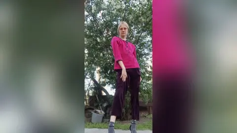 GeorgineAragan stands outdoors in a bright pink long-sleeve top and dark maroon pants against a backdrop of green trees.
