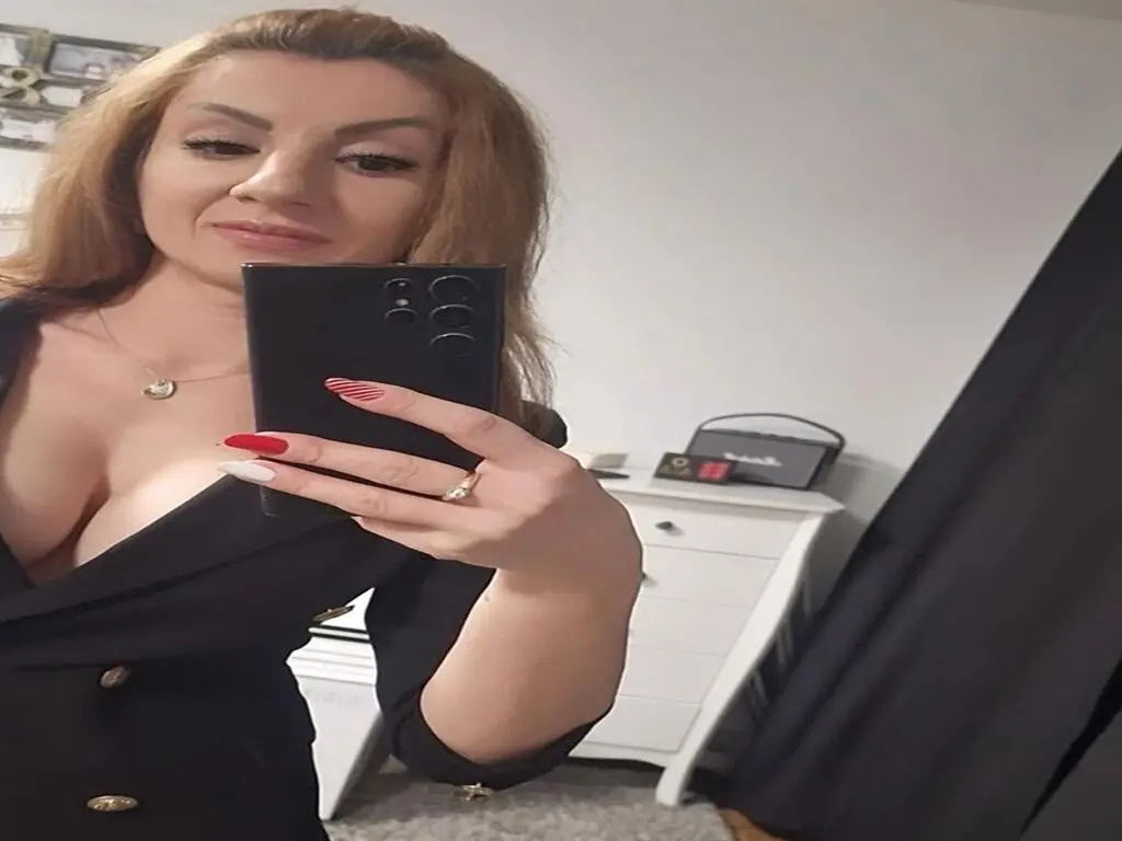 Smiling woman in a black low-cut dress taking a mirror selfie in a bright bedroom setting.