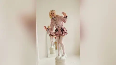 LynGoodhart smiles while posing in a pink ruffled dress and white lace stockings in a bright room.