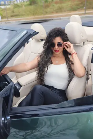 Smiling woman in a white tank top and sunglasses sitting in a luxury convertible car on a sunny day.