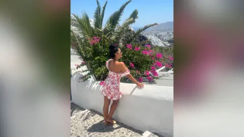 MissyJolie stands outdoors against a white wall wearing a short white and red floral sundress with off-the-shoulder sleeves.