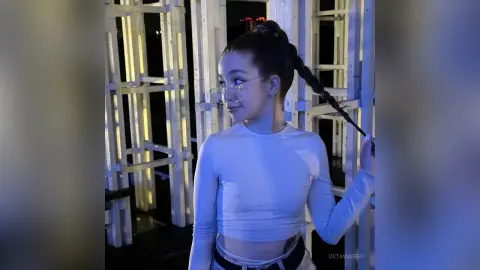 Young woman in a white long-sleeve crop top with a long braid posing in a modern blue-lit studio setting.