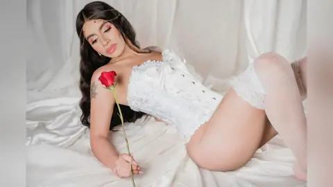 Woman in white lace lingerie holding a red rose while reclining on white silk sheets in a romantic mood.
