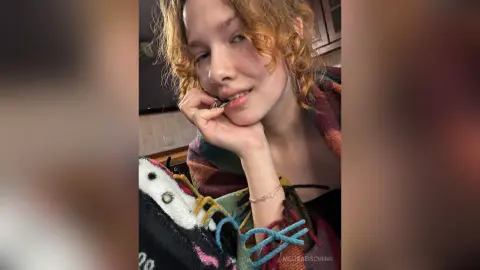 Smiling young woman with curly red hair wearing a cozy colorful patterned sweater in a bright indoor setting.
