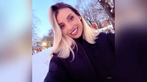 Smiling blonde woman in a black winter coat enjoying a sunny day in a snowy park.