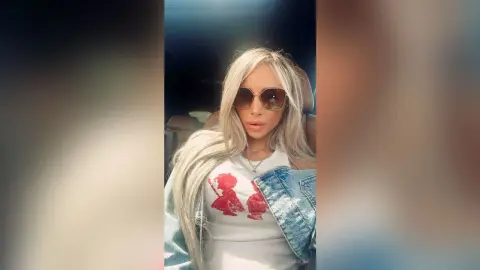 Blonde woman in a graphic white tank top and denim jacket wearing sunglasses inside a car.