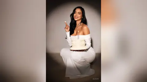 Alison smiles while wearing a white off-the-shoulder dress and holding a small white cake.