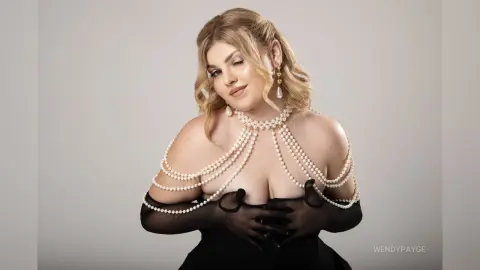 Elegant woman in a black dress with pearl body jewelry posing glamorously against a neutral studio background.