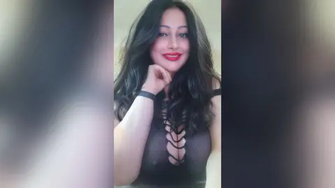 Smiling woman with long dark hair wearing a black lace cutout bodysuit in a bright indoor setting.