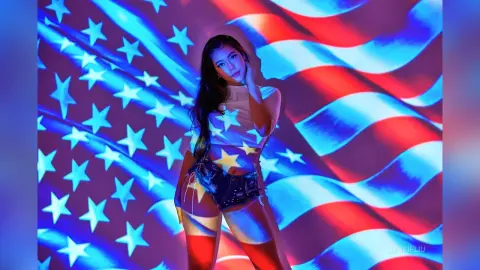 JessieLiu poses in front of a waving American flag, wearing a white tank top and denim shorts under blue and red lighting.