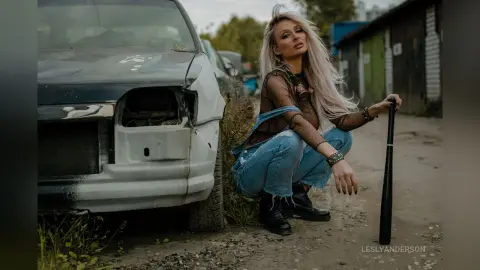 LeslyAnderson squats next to a white car, wearing a sheer black mesh top and blue denim jeans.