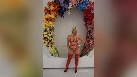 Smiling blonde woman in a red lace lingerie set and stockings posing against a colorful floral arch backdrop.