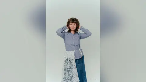 Smiling woman in a blue button-down shirt and white lace skirt posing against a plain white background.