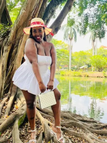 Smiling woman in a white sundress and straw hat holding a cream handbag by a tropical lake.