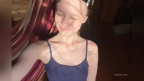 AmberLune smiles while wearing a blue tank top, with sunlight casting shadows across her face and chest.